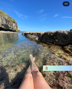 A blue sky above large rocks with a water inlet. Female legs in the foreground. 